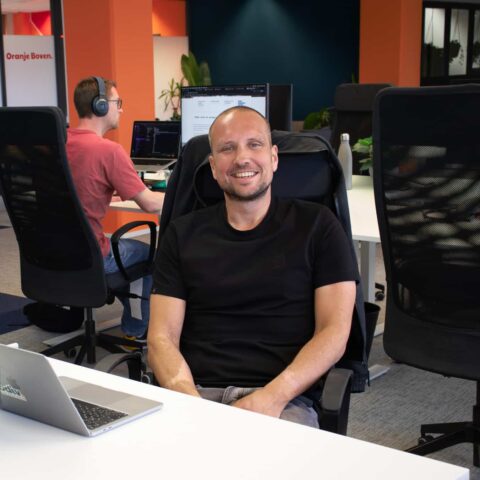 Een collega zit voor de computer en lacht naar de camera.